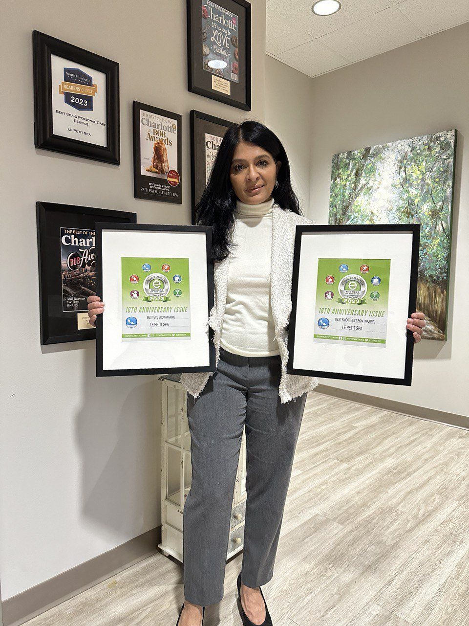 Priti holding two 2023 Elevate Lifestyle certificates in front of the framed award wall at Le Petit Spa