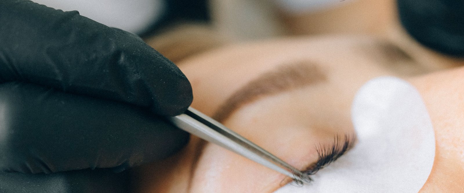 A gloved hand isolating a single lash with tweezers over a shaped eye pad.
